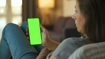 Woman at home lying on a couch and using smartphone with green mock-up screen in vertical mode. Girl browsing Internet, watching content, videos, blogs. POV. video