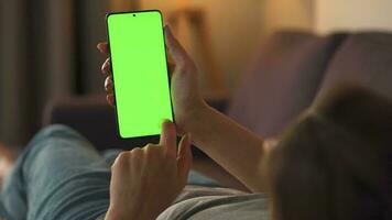 Woman at home lying on a couch and using smartphone with green mock-up screen in vertical mode. Girl browsing Internet, watching content, videos, blogs. POV. video