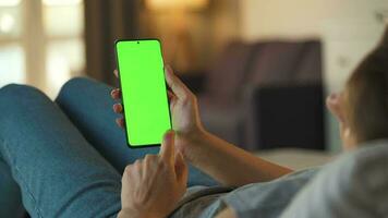 Woman at home lying on a couch and using smartphone with green mock-up screen in vertical mode. Girl browsing Internet, watching content, videos, blogs. POV. video