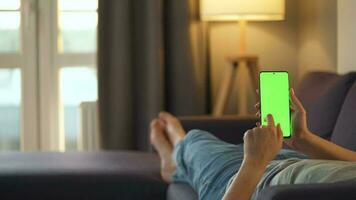 Woman at home lying on a couch and using smartphone with green mock-up screen in vertical mode. Girl browsing Internet, watching content, videos, blogs. POV. video