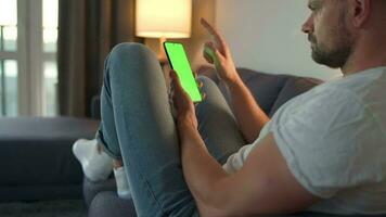 Man at home lying on a couch and using smartphone with green mock-up screen in vertical mode. He browsing Internet, watching content, videos, blogs. video