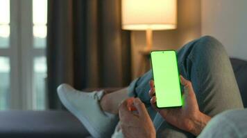 Man at home lying on a couch and using smartphone with green mock-up screen in vertical mode. He browsing Internet, watching content, videos, blogs. video