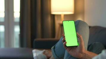 homem às casa deitado em uma sofá e usando Smartphone com verde brincar tela dentro vertical modo. ele navegando Internet, assistindo contente, vídeos, blogs. video