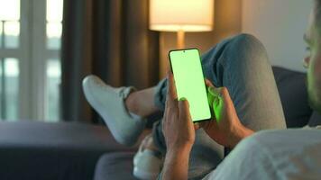 Man at home lying on a couch and using smartphone with green mock-up screen in vertical mode. He browsing Internet, watching content, videos, blogs. video