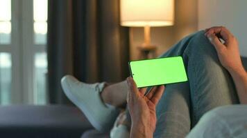 Man at home lying on a couch and using smartphone with green mock-up screen in vertical mode. He browsing Internet, watching content, videos, blogs. video