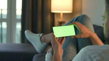 Man at home lying on a couch and using smartphone with green mock-up screen in vertical mode. He browsing Internet, watching content, videos, blogs. video