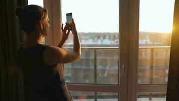 Frau steht in der Nähe von das Fenster und nimmt Bilder von das Rahmen Sonne auf ein Smartphone video