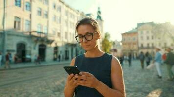 mulher caminhando baixa a velho rua e usando Smartphone às pôr do sol. comunicação, social redes, viagem conceito video