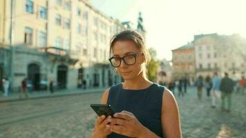 mulher caminhando baixa a velho rua e usando Smartphone às pôr do sol. comunicação, social redes, viagem conceito video