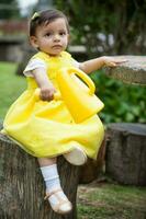 dulce uno año antiguo bebé niña vestido en amarillo riego el plantas a el jardín foto