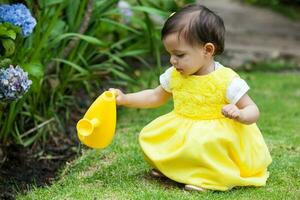 dulce uno año antiguo bebé niña vestido en amarillo riego el plantas a el jardín foto