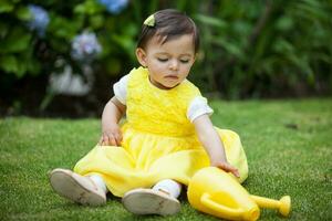 dulce uno año antiguo bebé niña vestido en amarillo riego el plantas a el jardín foto