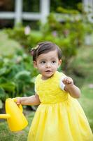 dulce uno año antiguo bebé niña vestido en amarillo riego el plantas a el jardín foto