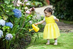 dulce uno año antiguo bebé niña vestido en amarillo riego el plantas a el jardín foto