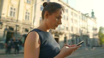 mulher caminhando baixa a velho rua e usando Smartphone às pôr do sol. comunicação, social redes, viagem conceito video