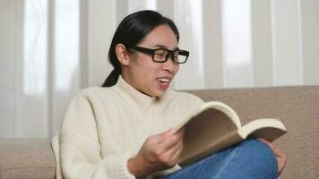 contento asiático mujer sentado en el sofá y leyendo un libro adentro. retrato de mujer en blanco suéter leyendo un libro en sofá a hogar. estilo de vida, gratis hora y relajación. video