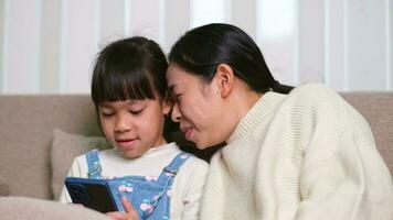 Young mother and her little daughter are using smartphone and smiling while sitting on sofa at home. Cheerful little girl using smartphone while relaxing with mom at home. Kids and Technology video