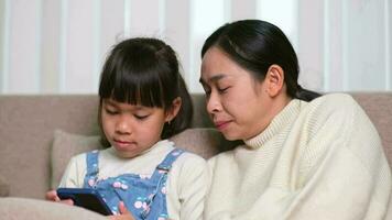 Jeune mère et sa peu fille sont en utilisant téléphone intelligent et souriant tandis que séance sur canapé à maison. de bonne humeur peu fille en utilisant téléphone intelligent tandis que relaxant avec maman à maison. des gamins et La technologie video