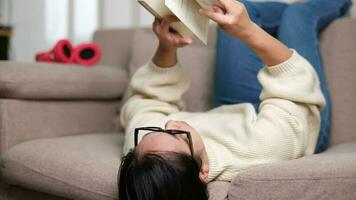 feliz ásia mulher deitado em a sofá e lendo uma livro dentro de casa. estilo de vida, livre Tempo e relaxamento. video