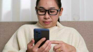 Happy young woman using mobile phone while sitting on sofa at home. Attractive woman holding smartphone checking social media, playing games, online shopping relaxing on sofa. video