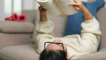 content asiatique femme mensonge sur le canapé et en train de lire une livre à l'intérieur. mode de vie, gratuit temps et relaxation. video