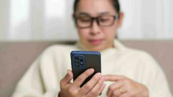 Happy young woman using mobile phone while sitting on sofa at home. Attractive woman holding smartphone checking social media, playing games, online shopping relaxing on sofa. video