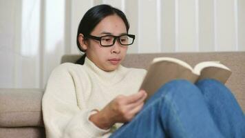 contento asiático mujer sentado en el sofá y leyendo un libro adentro. retrato de mujer en blanco suéter leyendo un libro en sofá a hogar. estilo de vida, gratis hora y relajación. video