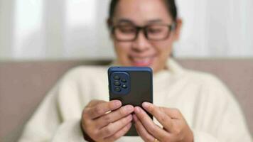 Happy young woman using mobile phone while sitting on sofa at home. Attractive woman holding smartphone checking social media, playing games, online shopping relaxing on sofa. video