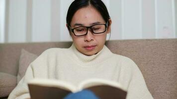 Happy Asian woman sitting on the sofa and reading a book indoors. Portrait of woman in white sweater reading a book on sofa at home. Lifestyle, free time and relaxation. video