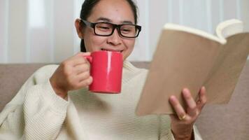 feliz jovem mulher segurando uma copo do café e lendo uma livro em a sofá depois de acordando acima dentro a manhã às lar. estilo de vida, livre Tempo e relaxamento. video