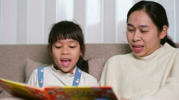 mère en train de lire une livre avec sa fille sur le canapé à maison. content mère en train de lire une livre, relaxant, étreindre, récit marrant histoires à les enfants et dépenses temps ensemble à maison. video