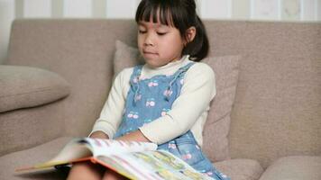 contento pequeño niña leyendo un libro adentro. linda pequeño niña en casual ropa sentado en el sofá leyendo un libro. video