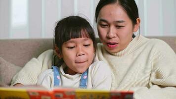 Mother reading a book with her daughter on the sofa at home. Happy mother reading a book, relaxing, hugging, telling funny stories to children and spending time together at home. video