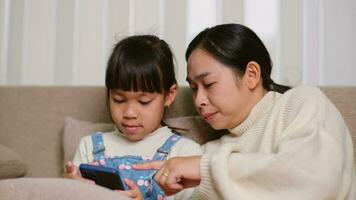 joven madre y su pequeño hija son utilizando teléfono inteligente y sonriente mientras sentado en sofá a hogar. alegre pequeño niña utilizando teléfono inteligente mientras relajante con mamá a hogar. niños y tecnología video