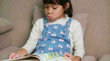 feliz pequeno menina lendo uma livro dentro de casa. fofa pequeno menina dentro casual roupas sentado em a sofá lendo uma livro. video
