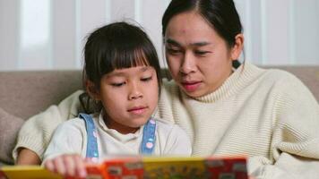 mère en train de lire une livre avec sa fille sur le canapé à maison. content mère en train de lire une livre, relaxant, étreindre, récit marrant histoires à les enfants et dépenses temps ensemble à maison. video