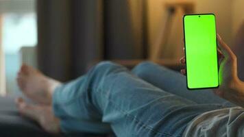 Woman at home lying on a couch and using smartphone with green mock-up screen in vertical mode. Girl browsing Internet, watching content, videos, blogs. POV. video