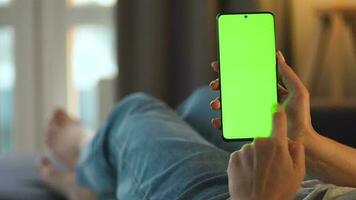 vrouw Bij huis aan het liegen Aan een bankstel en gebruik makend van smartphone met groen mock-up scherm in verticaal modus. meisje browsen internetten, aan het kijken inhoud, videos, bloggen. gezichtspunt video