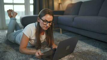 vrouw is aan het liegen Aan de verdieping en en maakt een online aankoop gebruik makend van een credit kaart en laptop. online winkelen, levensstijl video