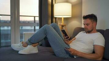 Man with glasses lying on the couch in a cozy room and using smartphone for surfing internet or chatting in messengers video