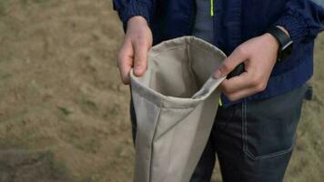 un niño en un azul chaqueta y pantalones soportes en el arena y sostiene un bolso de ropa foto