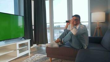 Man at home lying on a couch and watching TV with green mock-up screen video