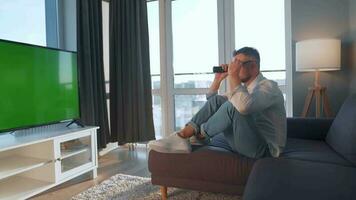 Man at home lying on a couch and watching TV with green mock-up screen video