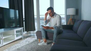 homme avec des lunettes séance sur le canapé dans une confortable chambre, Parlant sur une téléphone intelligent et en utilisant portable. éloigné travail concept video