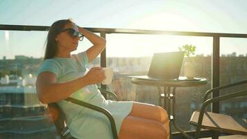 Woman drinks coffee and rests during a break on the balcony against the the setting sun. Remote work concept video