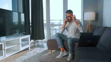 hombre con lentes sentado en el sofá en un acogedor habitación, Hablando en un teléfono inteligente y utilizando ordenador portátil. remoto trabajo concepto video