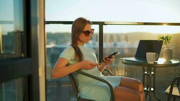 femme séance sur le balcon contre le toile de fond de le réglage Soleil et fait du un en ligne achat en utilisant une crédit carte video