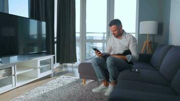 hombre con lentes sentado en el sofá en un acogedor habitación, Hablando en un teléfono inteligente y utilizando ordenador portátil. remoto trabajo concepto video