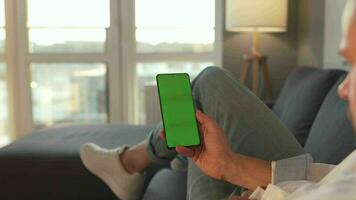 Man at home lying on a sofa and using smartphone with green mock-up screen in vertical mode. Guy browsing Internet video