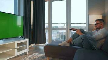 Man at home lying on a couch and watching TV with green mock-up screen video
